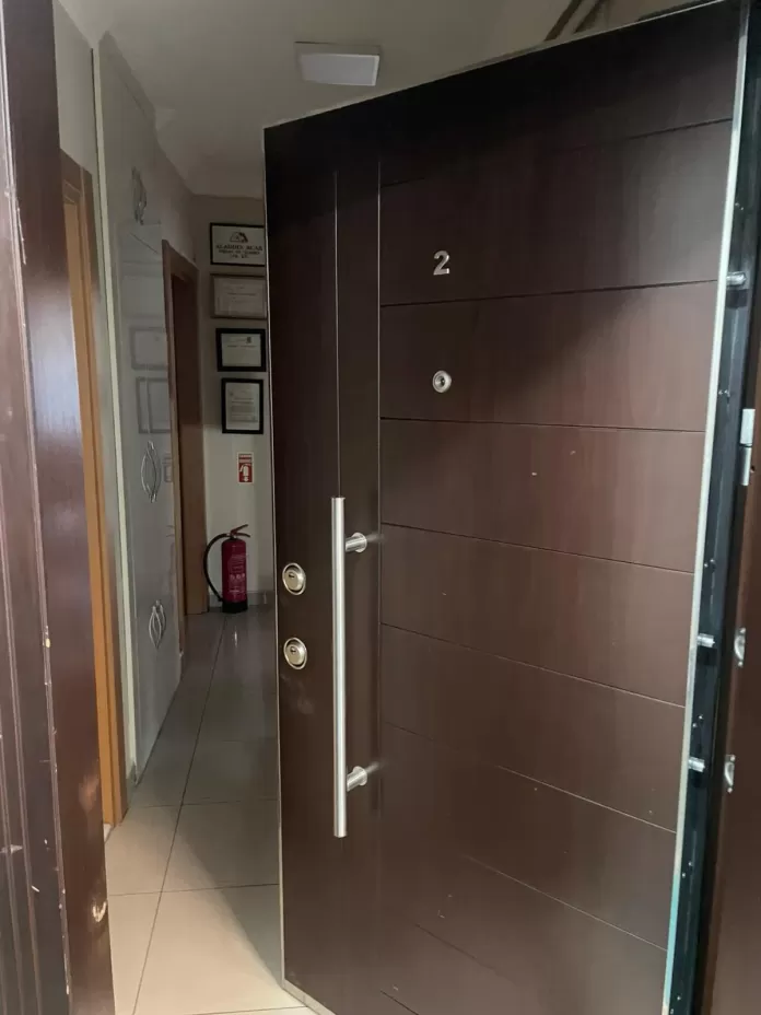 Apartman koridorunda koyu kahverengi çelik daire kapısı; iki barel rozeti, boru kulp ve gözetleme deliği