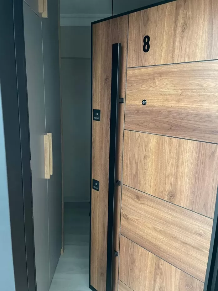 Montajı tamamlanmış ahşap desenli çelik daire kapısı: siyah kasa, uzun barel kulp, gözetleme deliği ve daire numarası